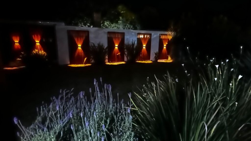 Vista nocturna de jardín con iluminación cálida bidireccional instalada sobre paneles de madera en el muro perimetral.
