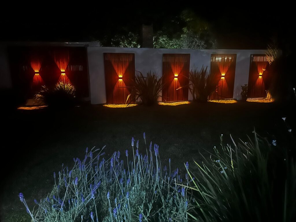 Primer plano de aplique de luz encendido sobre panel de madera exterior, creando un ambiente cálido en el jardín de noche.